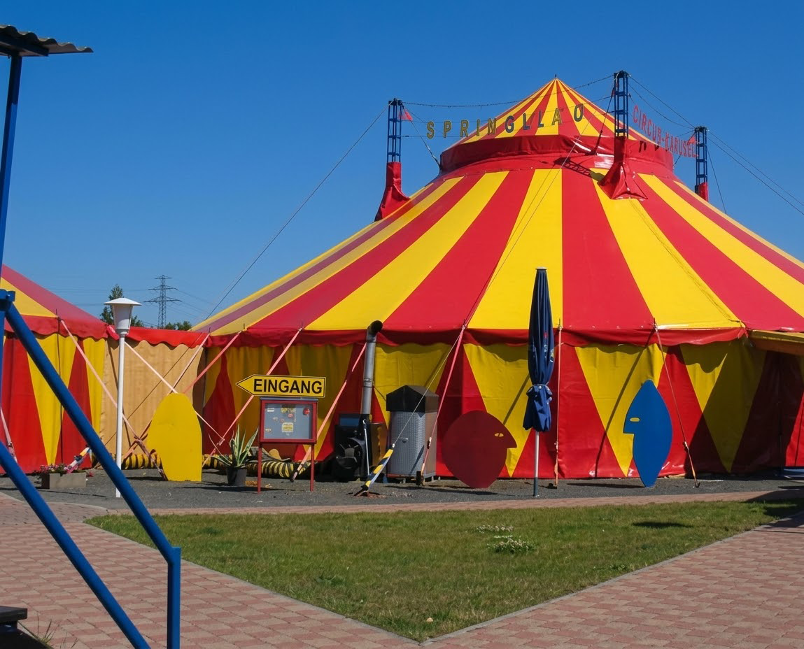 Rotes und gelbes Zirkuszelt vor blauem Himmel mit "Eingang"-Schild